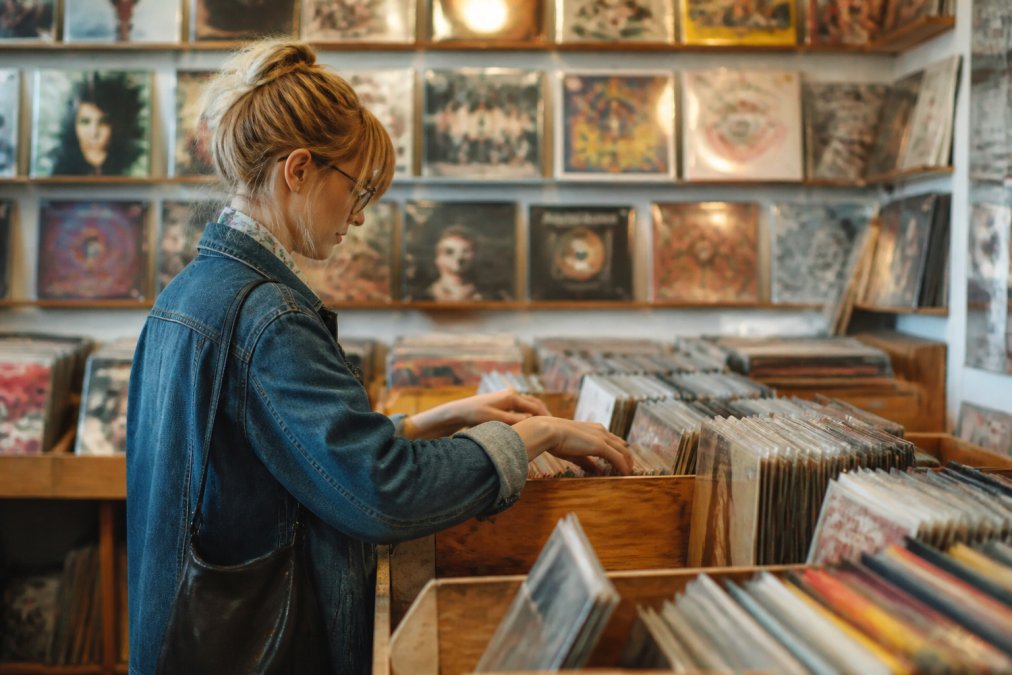 A person browsing through vinyl records in a cozy, well-lit independent record store, with wooden bins and colorful album covers visible