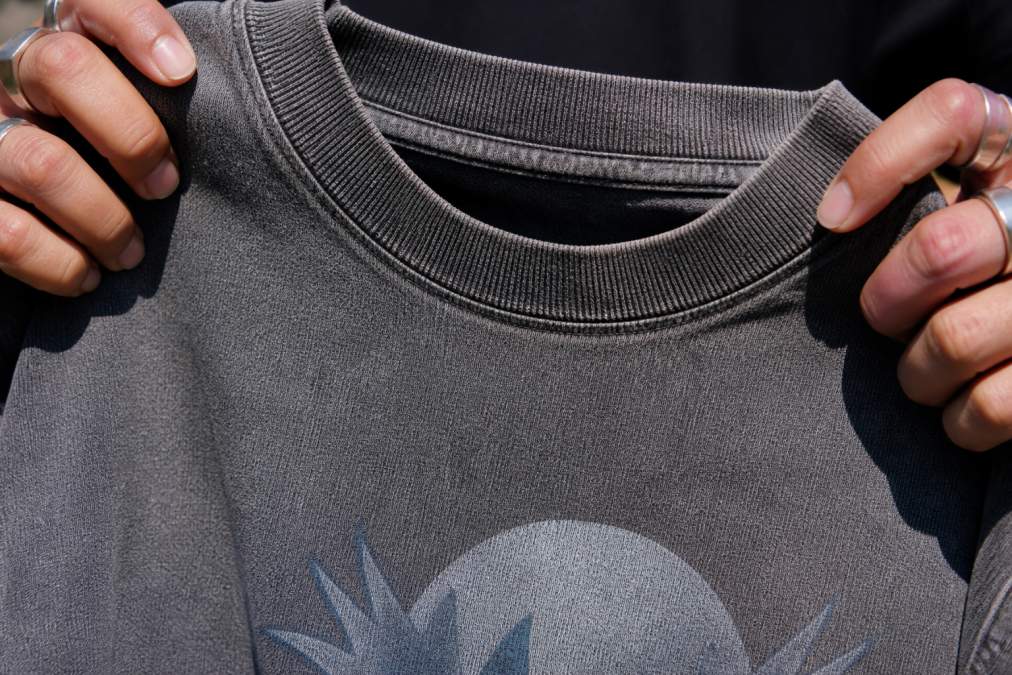 Close-up of hands holding a heavyweight cotton graphic tee against natural light, showing the thick fabric texture, snow-washed fade, and stitched crew neck detail