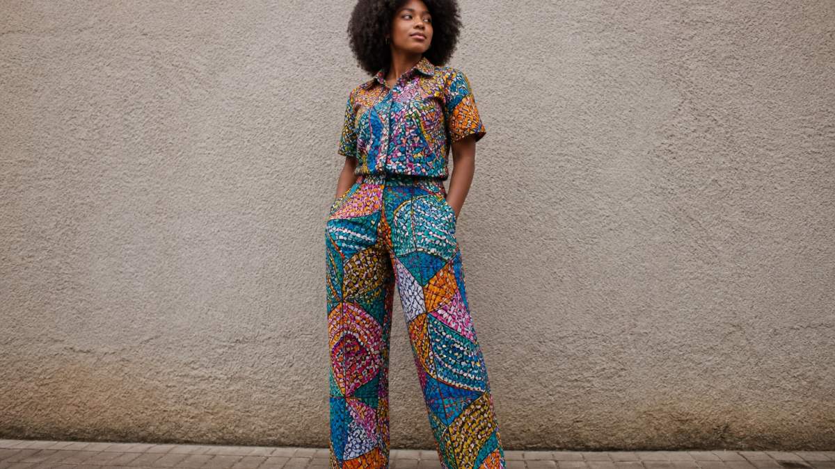Person wearing a vibrant multicoloured Ankara print co-ord set in natural daylight, standing confidently with hands in pockets, showing rich fabric texture and bold geometric patterns against a neutral background