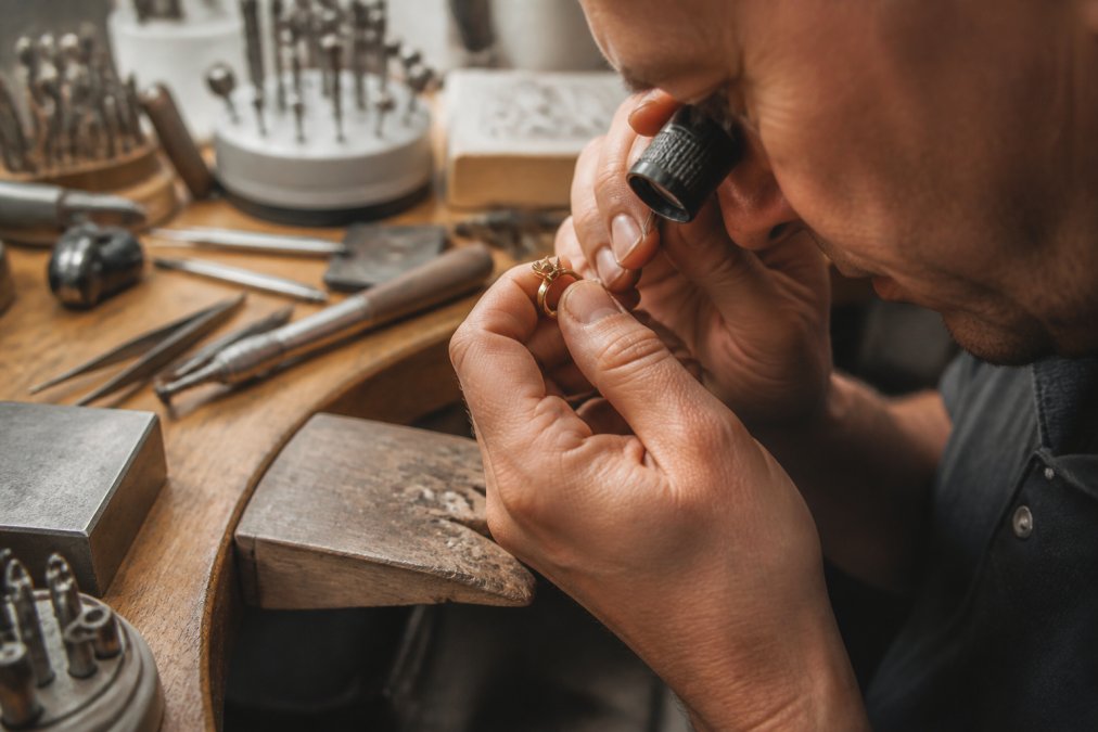 Close-up of a jeweler's hands examining a custom gold ring under a jeweler's loupe, inspecting stone setting and metal work details in a bright workshop setting