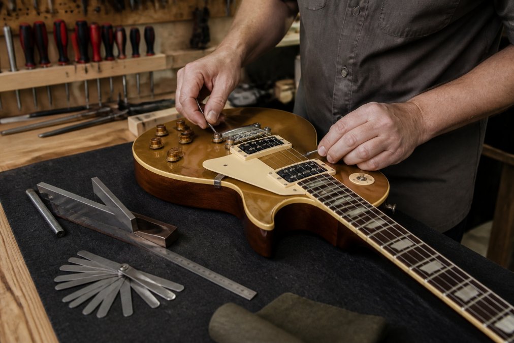 Luthier's hands adjusting the bridge and strings of a guitar on a workbench, with precision measurement tools, feeler gauges, and a straightedge visible, demonstrating professional setup work in a well-lit workshop