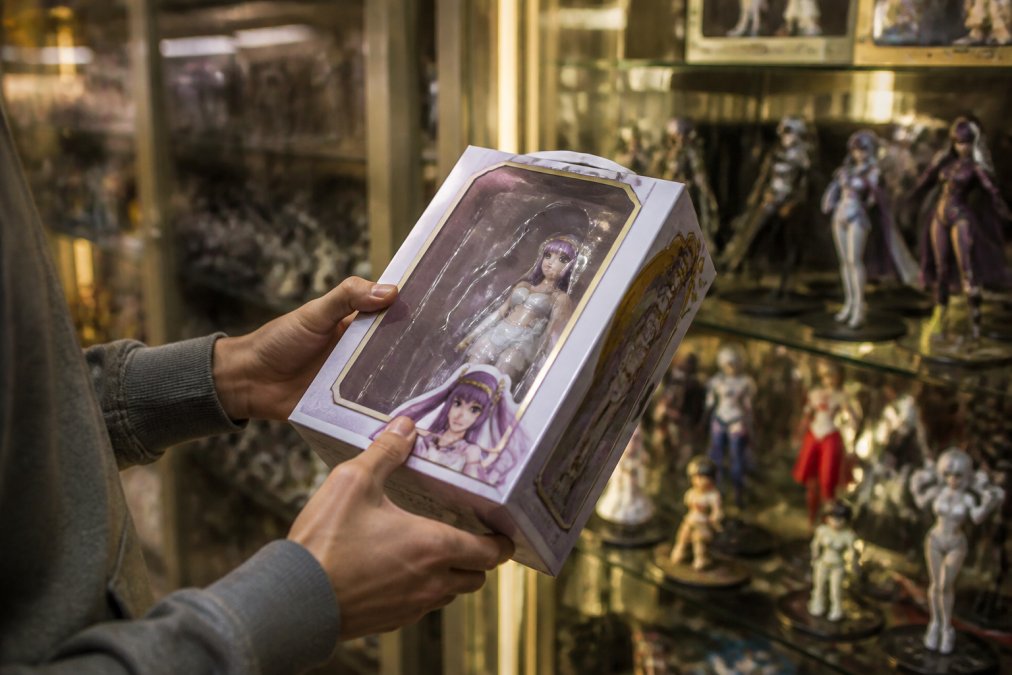 Close-up of a collector's hands carefully examining a rare anime figure still in its original box, with shelves of other collectibles blurred in the background, warm retail lighting
