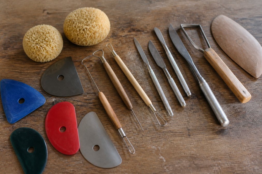 Flat lay of essential pottery tools arranged on a wooden work surface in natural studio lighting: polymer ribs in various colors, wire clay cutters, needle tools, natural sponges, and stainless steel trimming tools