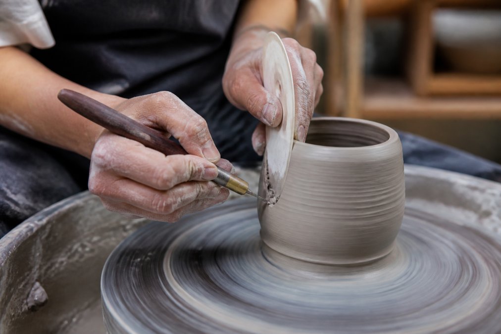 Close-up of hands holding a precision carving tool with hardwood handle and polymer rib while shaping clay on an active pottery wheel, with natural clay dust visible in soft studio lighting