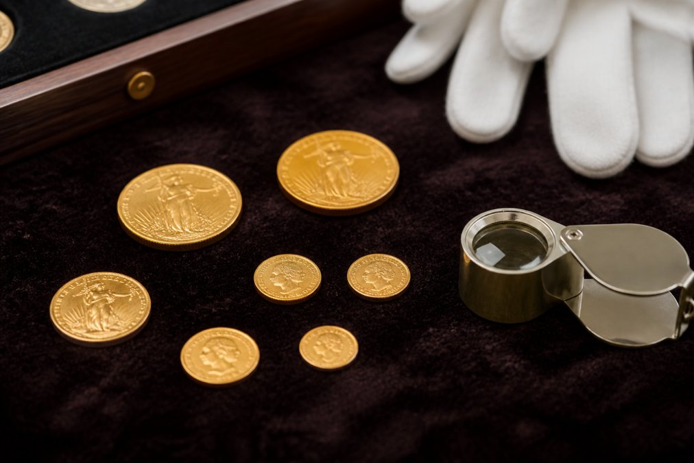 Close-up of several small gold coins of different sizes arranged on a dark velvet surface, with a jeweler's loupe and white cotton gloves nearby, warm natural lighting
