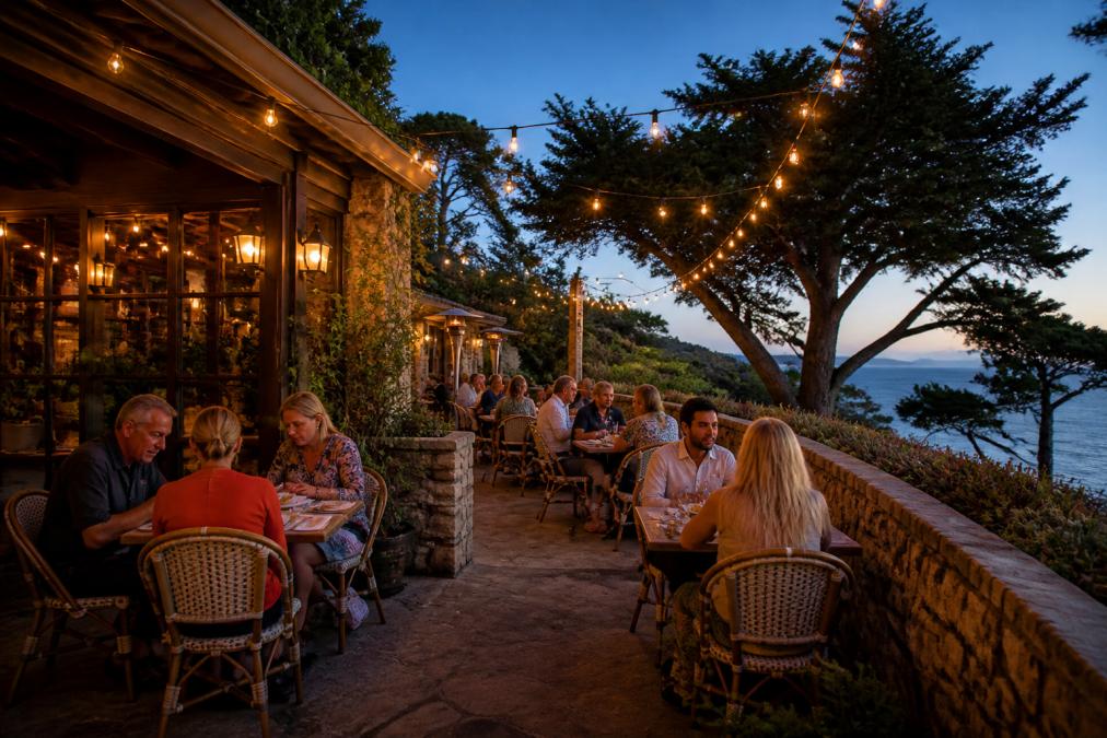 A warmly lit restaurant patio in Carmel-by-the-Sea at dusk, string lights overhead, diners at small tables, coastal foliage and a stone wall nearby