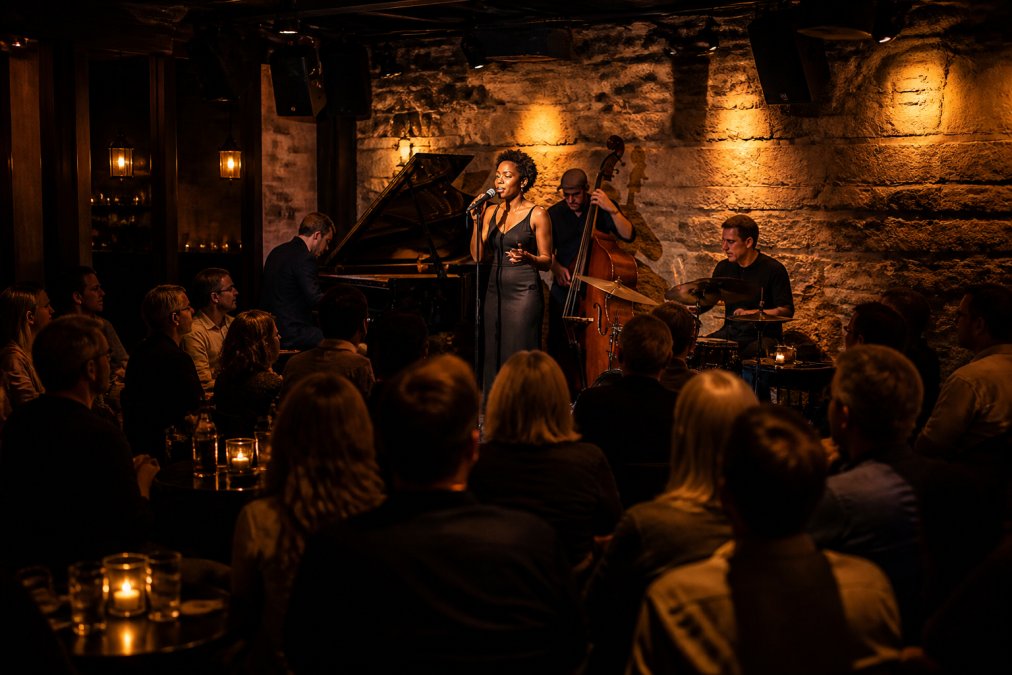A performer on stage in an intimate jazz club with warm amber lighting, close audience seating arranged around the stage, and a focused, attentive crowd