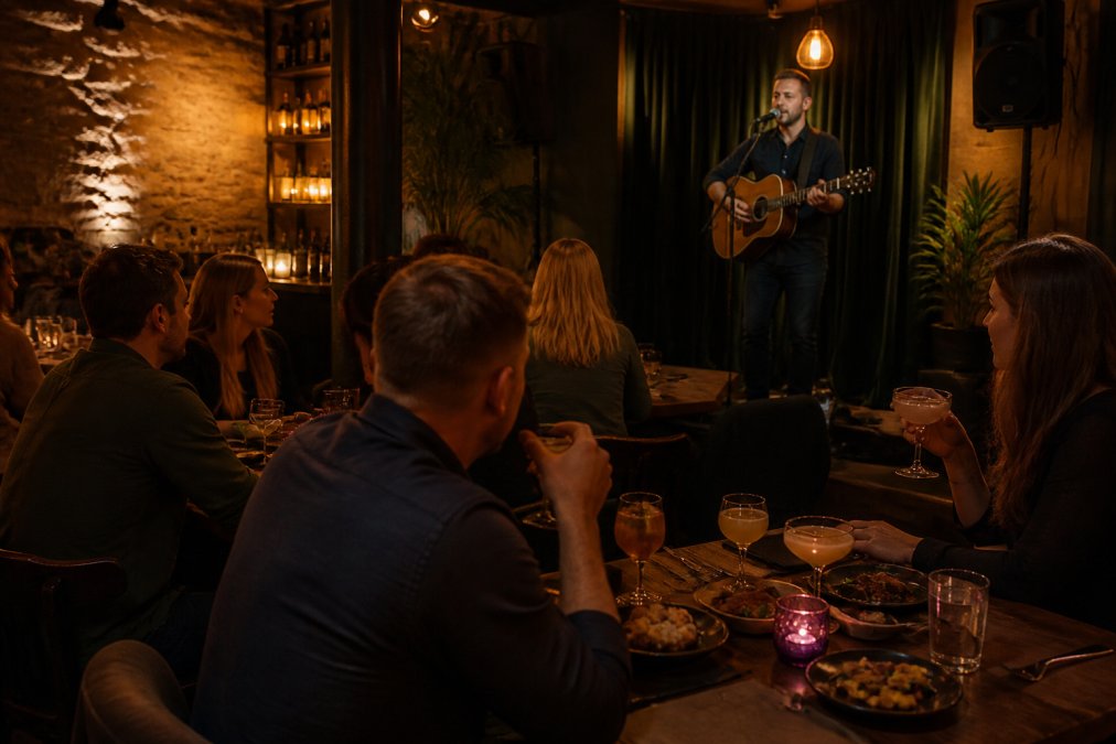 A restaurant bar scene with a live musician performing on a small stage in the background, diners seated at tables enjoying small plates and craft cocktails, warm ambient lighting creating an intimate atmosphere