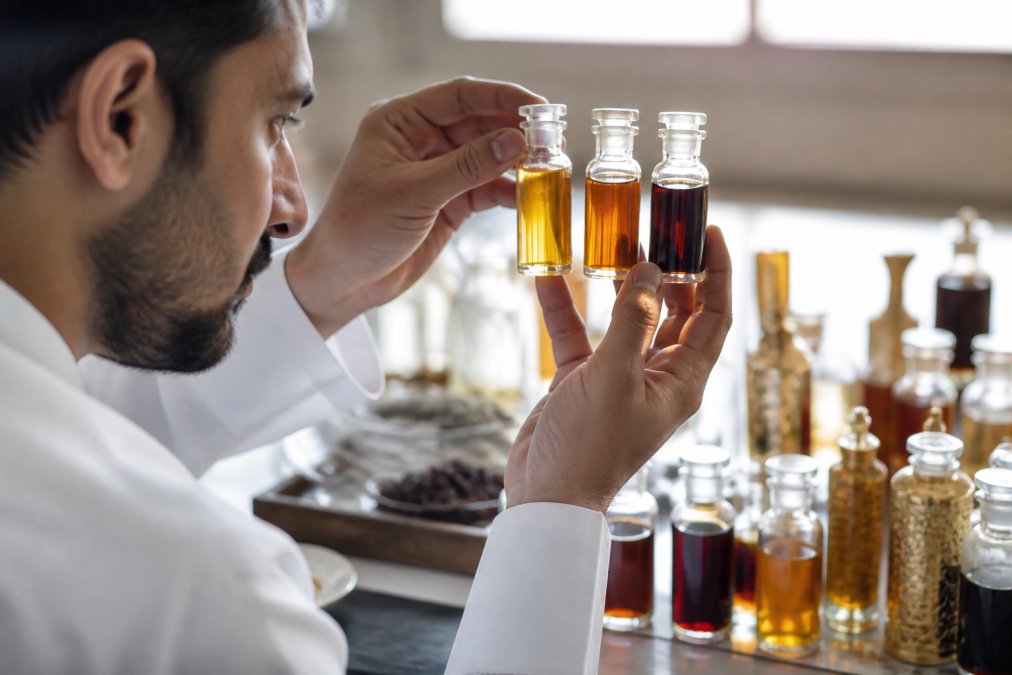 Professional examining and comparing bottles of premium oud attar oils against natural light, showing color clarity and viscosity differences in traditional glass vials