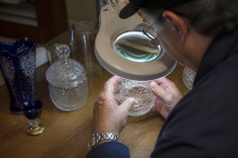 Collector examining vintage glassware item under bright magnifying lamp, hands carefully inspecting surface details and markings on wooden table