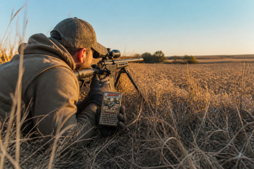 A hunter crouched low in tall grass using an electronic predator call device, scanning a field edge with a scoped rifle rested on a bipod, late afternoon golden light across the field