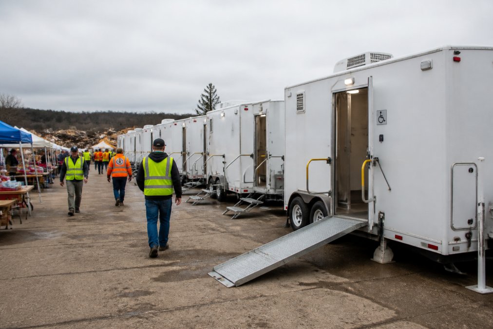 A row of clean, modern accessible shower trailers parked at a disaster relief staging area, with volunteers in safety vests walking between units and a clear ADA ramp visible on the nearest trailer