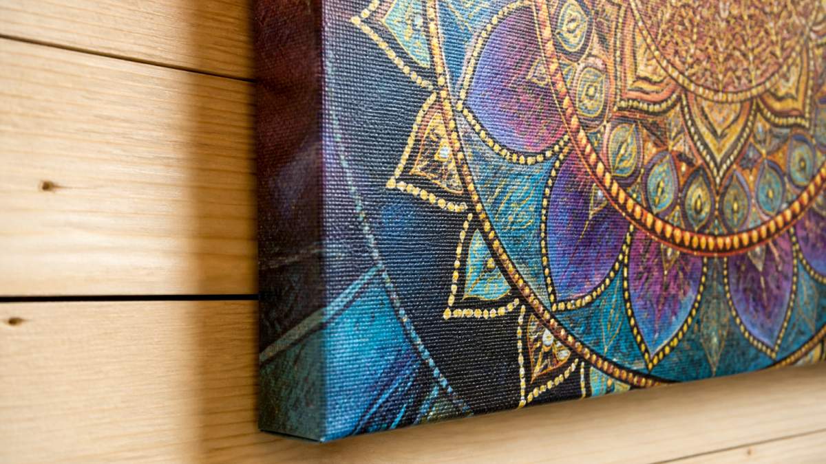 Close-up of a stretched canvas print displayed on a light wooden wall, showing fine texture detail of premium canvas material, rich color saturation in a mandala design, and neat edge wrapping where the image wraps around the wooden stretcher bars