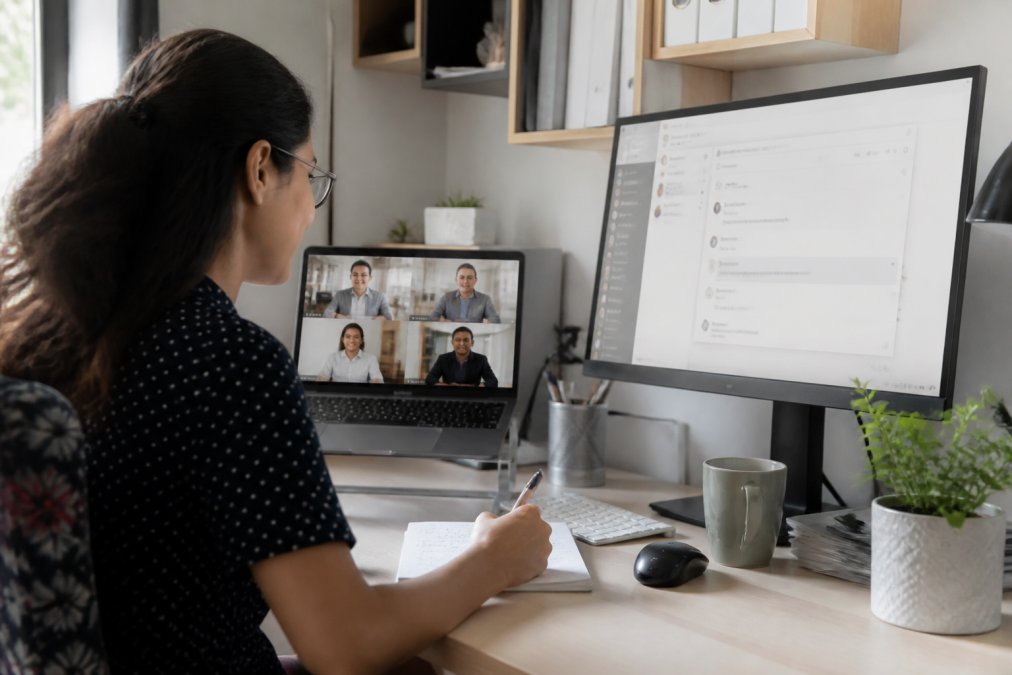 Team member using video conferencing on laptop with dual monitors in professional home office, screen showing secure meeting interface, encrypted messaging visible on secondary monitor, demonstrating secure remote collaboration setup