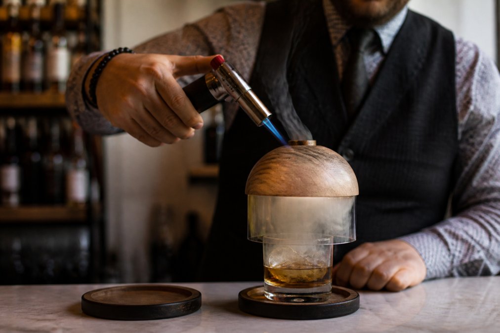 A butane torch ignites wood chips in a whiskey smoking set dome over a rocks glass.