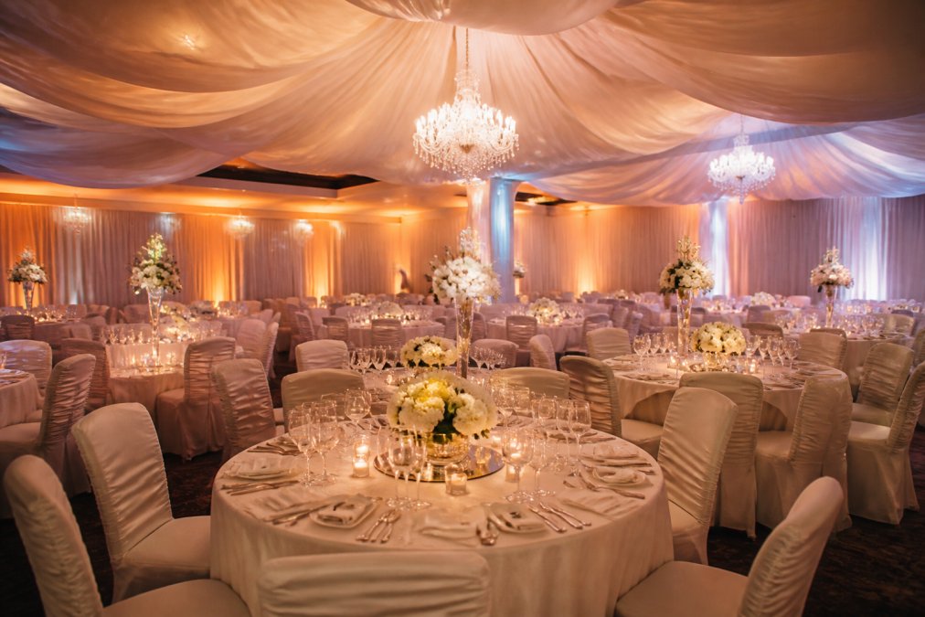 An elegant banquet hall interior with draped ceiling fabric, round tables with white linens, and warm up-lighting along the walls, set for a formal reception
