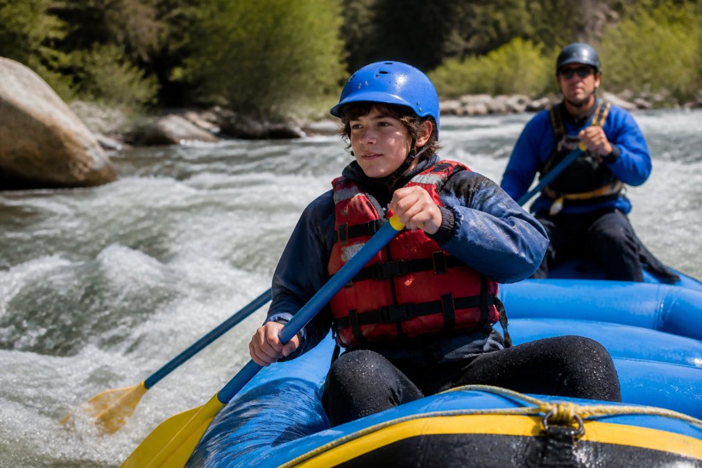 A teenage rafter wearing a properly fitted life jacket and helmet, paddling through Class II rapids with a professional guide visible in the background