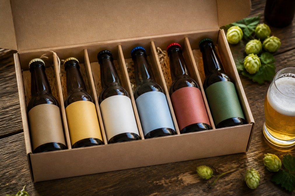 A close-up photograph of an open cardboard gift box containing six 330ml bottles of alcohol-free beer, styled on a rustic wooden table with hops and a glass of poured beer beside it
