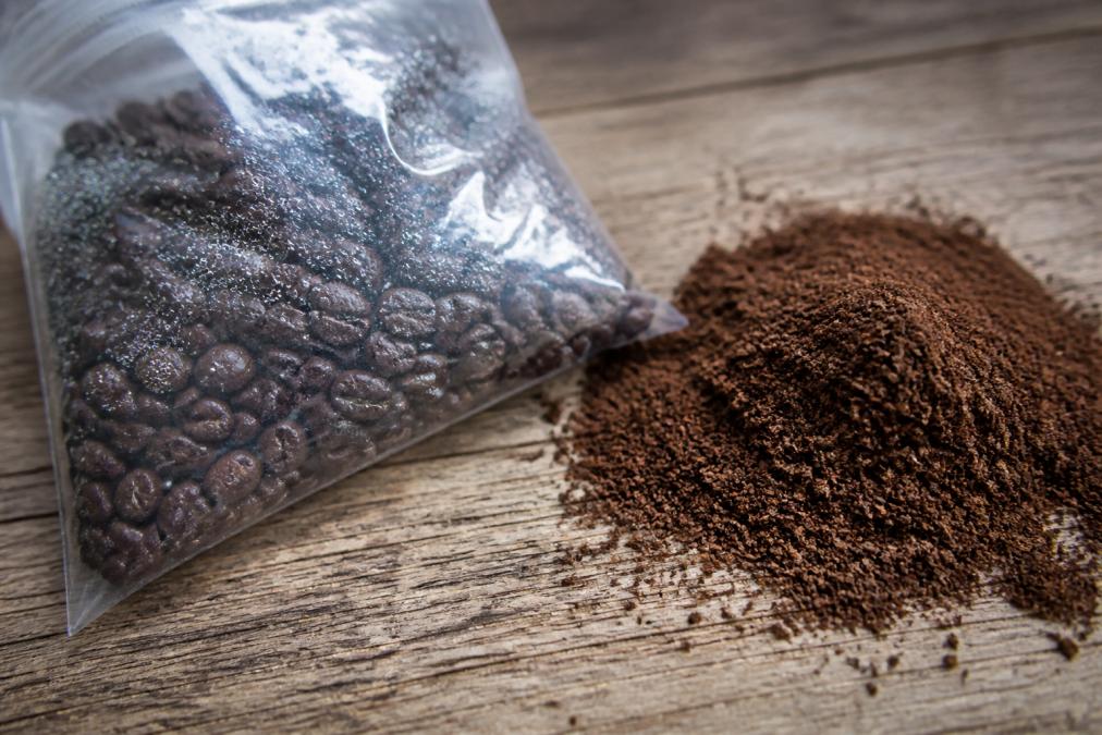 Close-up of whole coffee beans in a sealed transparent bag showing visible bloom and gas release, positioned next to a pile of pre-ground coffee displaying surface oxidation and browning, natural lighting highlighting the contrast