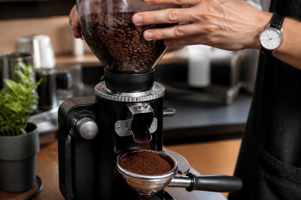 Hands positioning whole coffee beans into a burr grinder hopper, with freshly ground coffee falling into a filter basket below, showing the grinding mechanism in action and the resulting uniform ground texture