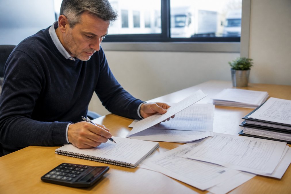 Fleet manager reviewing vehicle maintenance records and cost reports at a desk with multiple documents and a calculator, natural office lighting