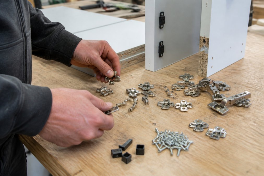 Professional joinery workshop workbench with assorted cabinet hardware components including hinges, confirmat screws, connectors, and metal fittings arranged for a kitchen cabinet project, with hands selecting pieces