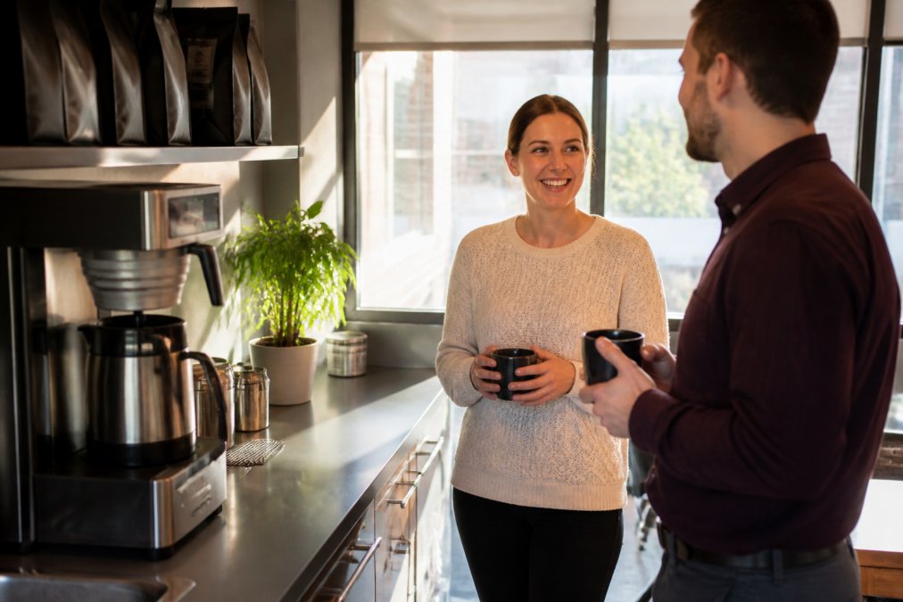 Office breakroom with a commercial coffee brewer on the counter, whole bean bags visible on a shelf, warm morning light, two colleagues filling mugs and chatting