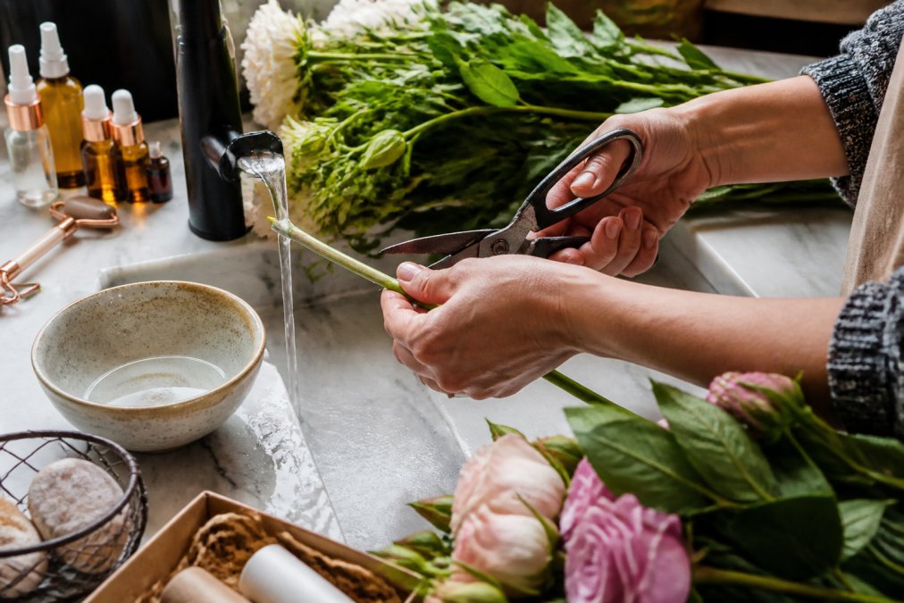 Close-up of hands conditioning flower stems under running water, trimming stems at a 45-degree angle with sharp floral scissors, with fresh cut flowers and conditioning supplies arranged on a clean countertop