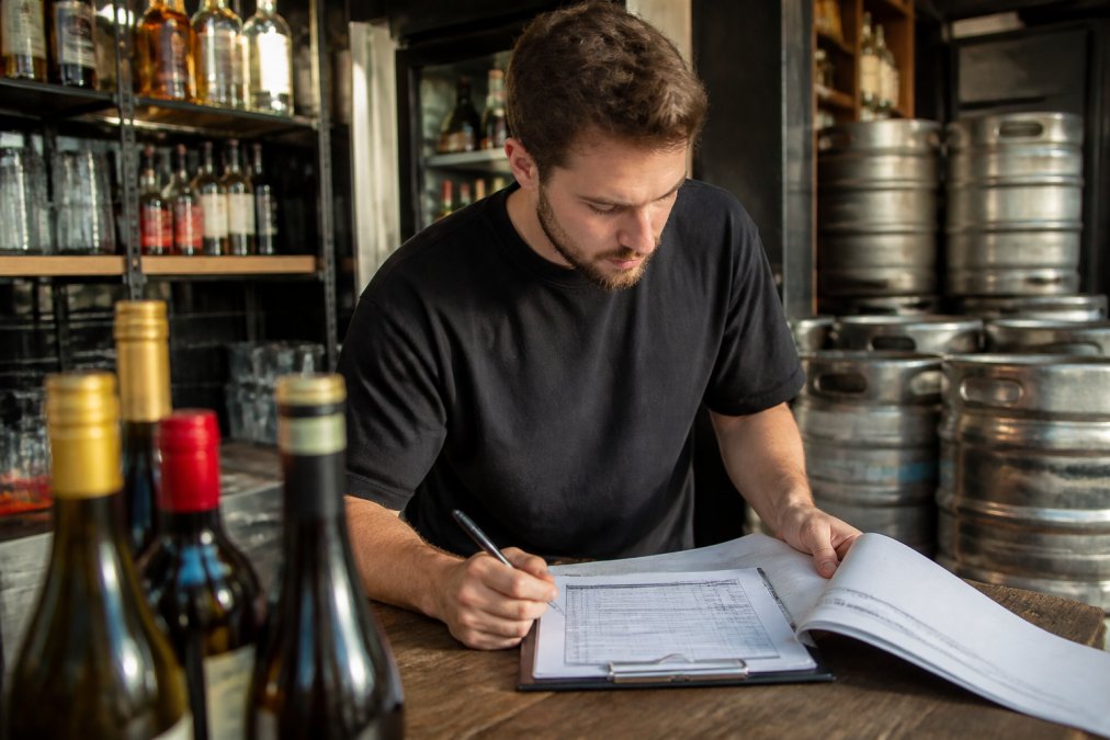 Bar manager reviewing supplier order forms and stock documentation at a hospitality venue counter, with bottles and kegs visible in background, natural afternoon light