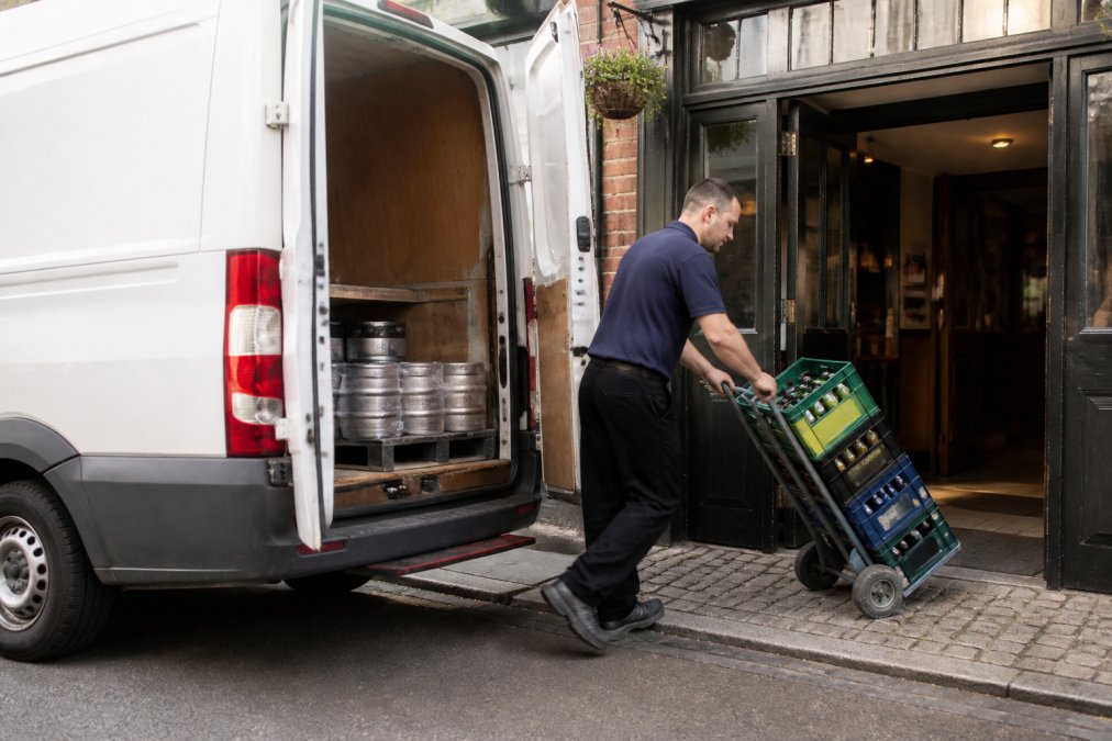 White van delivering crates of drinks to a pub entrance, driver unloading stock into venue, daytime lighting
