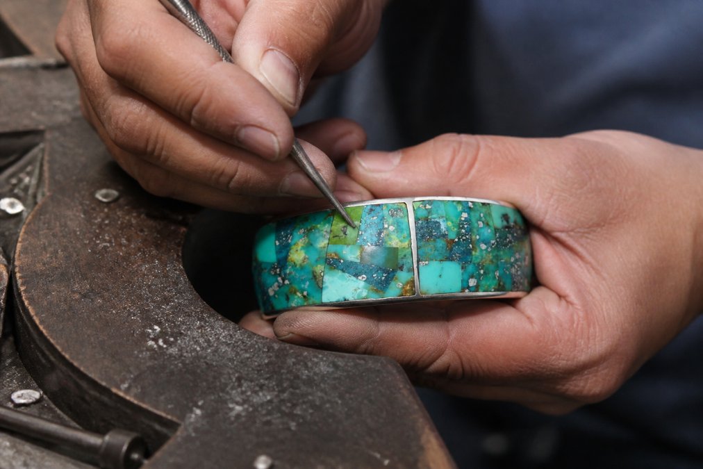 Close-up of artisan hands carefully setting turquoise stones into a sterling silver cuff using traditional inlay techniques, showing detailed silversmithing work on a jeweler's bench with natural lighting
