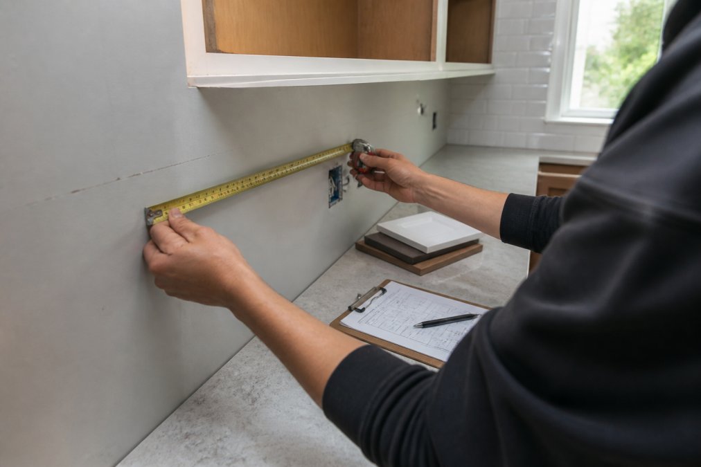 Close-up of hands using a metal tape measure on kitchen wall with cabinet space visible, clipboard with measurements and notes on countertop nearby