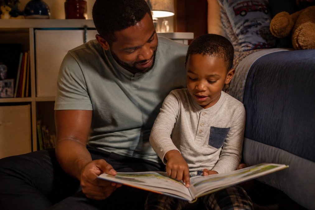 Adult and child sitting together with an open storybook, child pointing at illustrations while the adult smiles, warm home setting