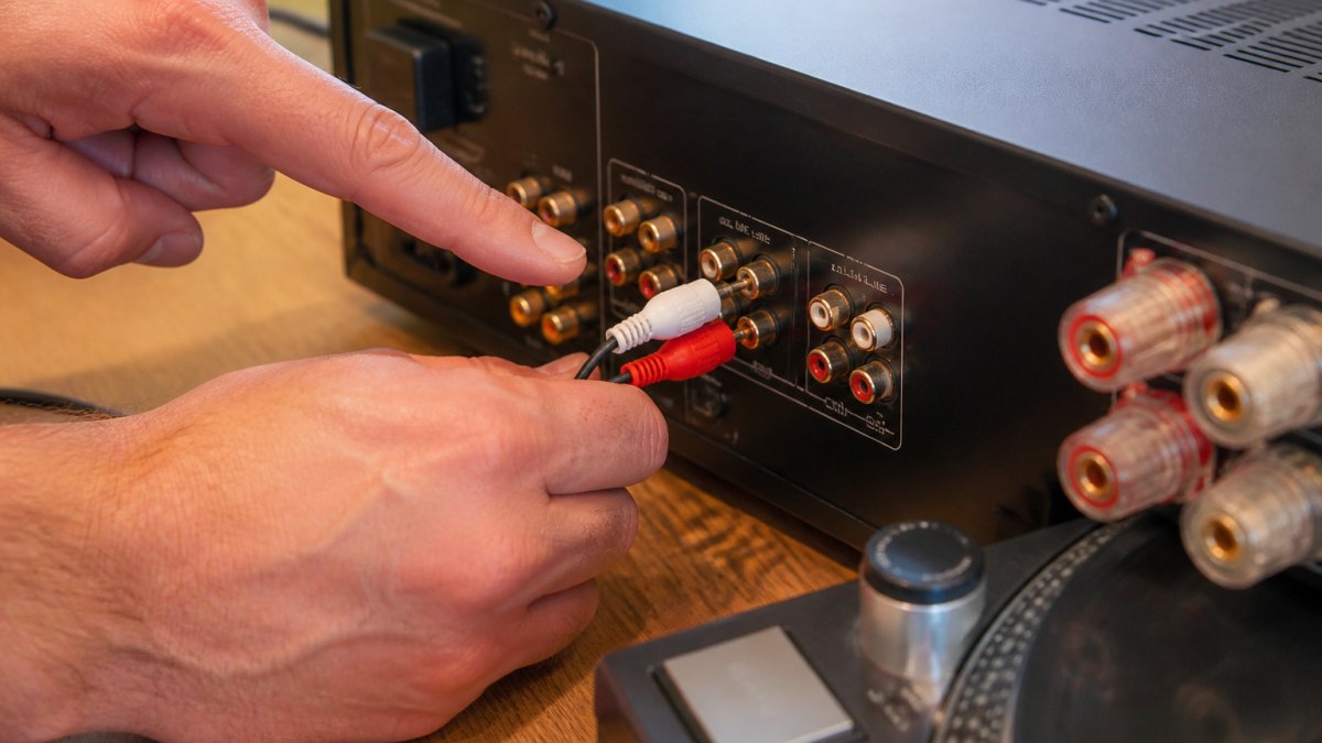 Close-up of turntable RCA cables colour-coded red and white being connected to an amplifier's phono input terminals, with a hand pointing to the labelled connectors