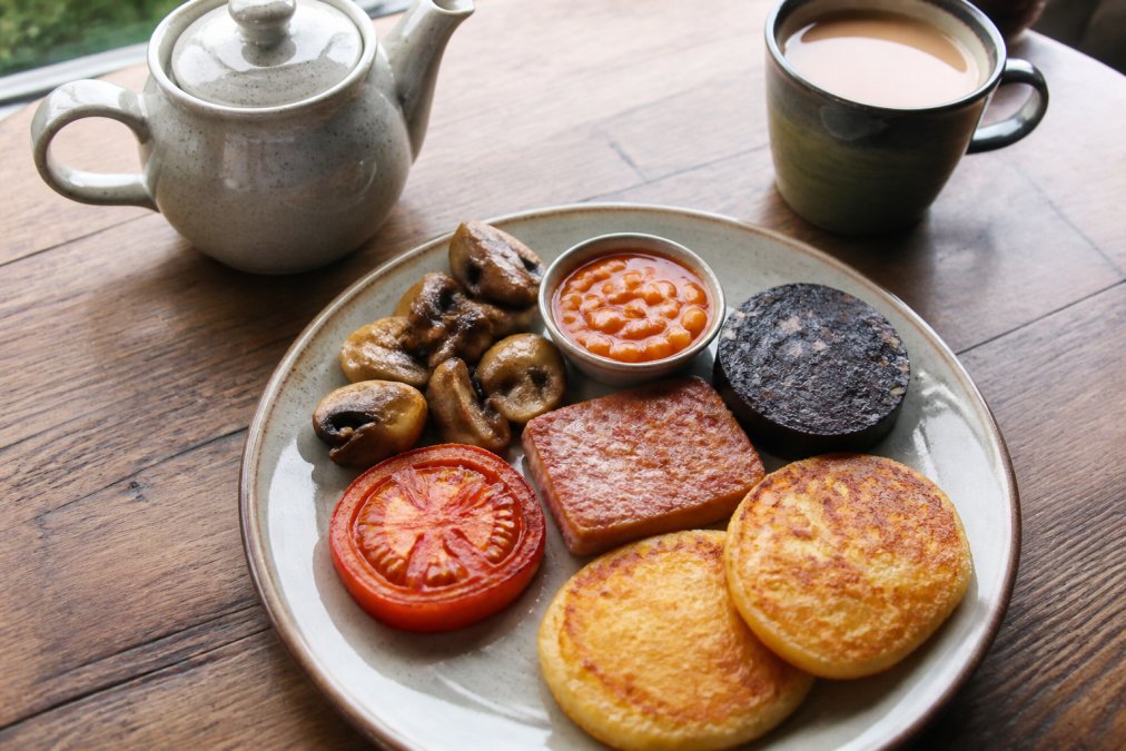 A close-up overhead shot of a full traditional Scottish breakfast plate on a rustic wooden table, featuring Lorne sausage, black pudding, tattie scones, grilled tomato, mushrooms, and a pot of tea, with soft morning light from a window