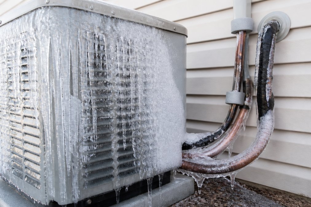 Close-up of thick ice buildup accumulated on an outdoor AC condenser unit and copper refrigerant lines on a residential home exterior, with frost visible in morning sunlight