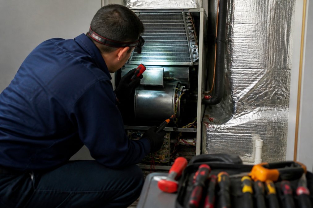 Licensed HVAC technician in uniform inspecting and examining an indoor air handler unit, checking the evaporator coil and blower motor area with a flashlight and diagnostic tools