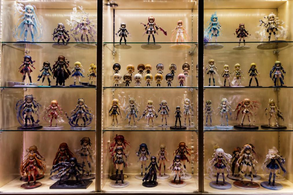 Carefully arranged anime figure collection on illuminated glass shelves displaying scale figures, Nendoroids, and PVC statues with varied poses and character designs, organized by height and theme against neutral wall tones with warm accent lighting