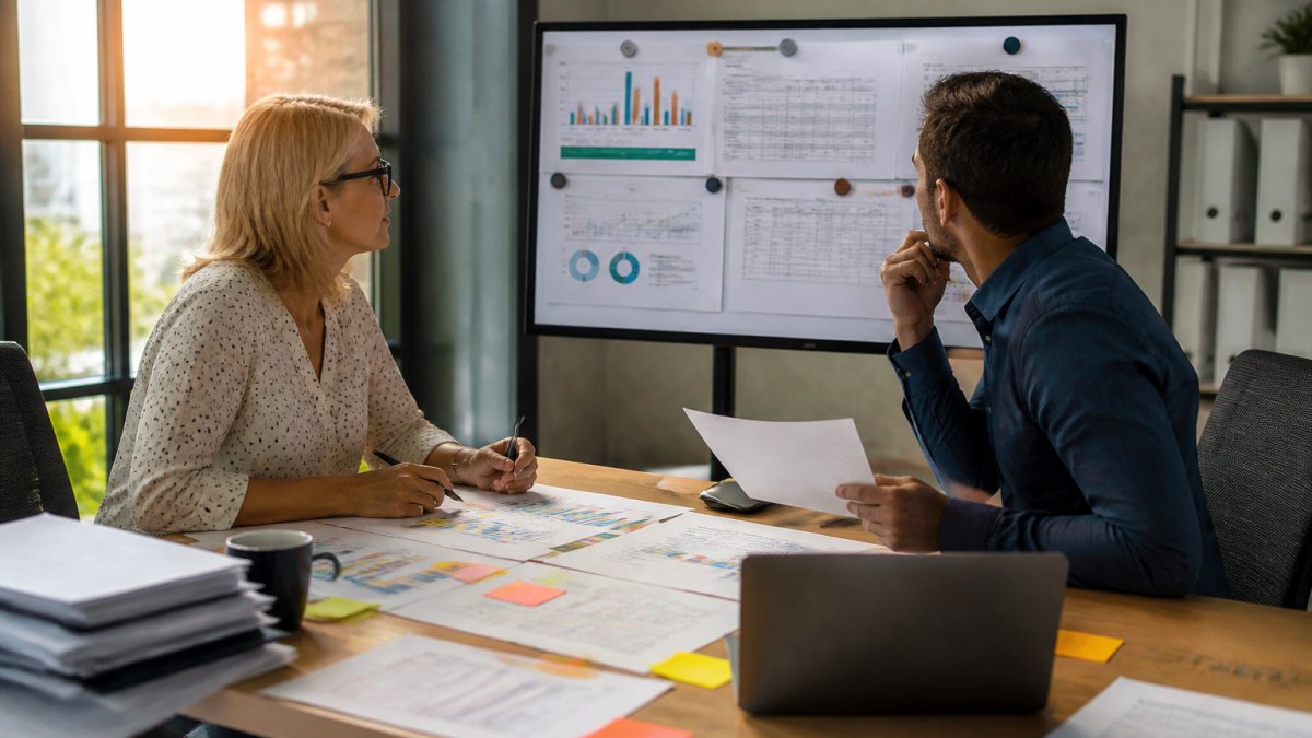 A sales manager and a data analyst reviewing forecast projections on a large monitor in a modern office, printed pipeline reports and sticky notes on the desk, warm afternoon light
