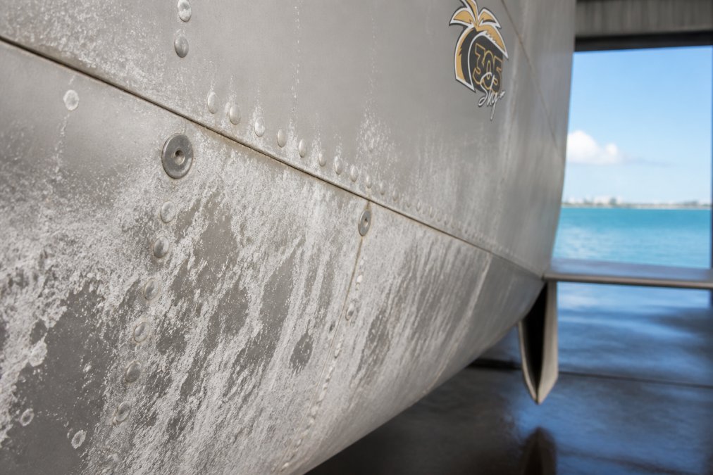 Close-up of aircraft fuselage showing white salt corrosion deposits and oxidation on aluminum skin near fasteners and seams in a coastal hangar environment with ocean visible in background