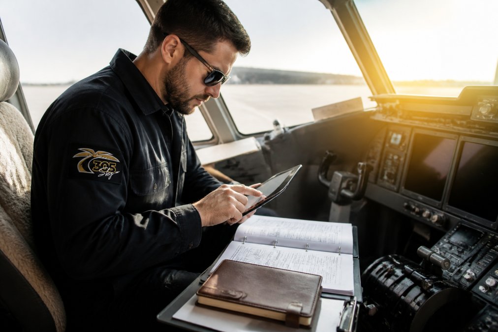 Aviation maintenance technician in uniform reviewing detailed maintenance logs on digital tablet in aircraft cockpit, with printed maintenance manual and leather-bound logbook visible on clipboard nearby, natural sunlight streaming through cockpit window