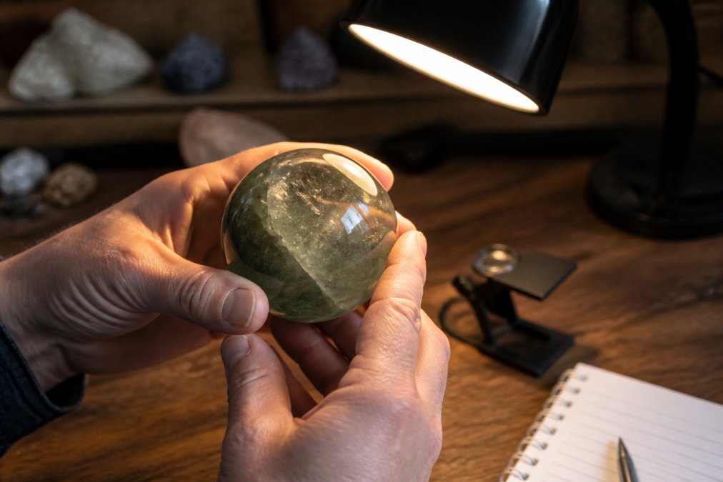 Close-up of a person's hands carefully examining a polished crystal sphere under a warm lamp, with a loupe and notebook on a wooden table in the background