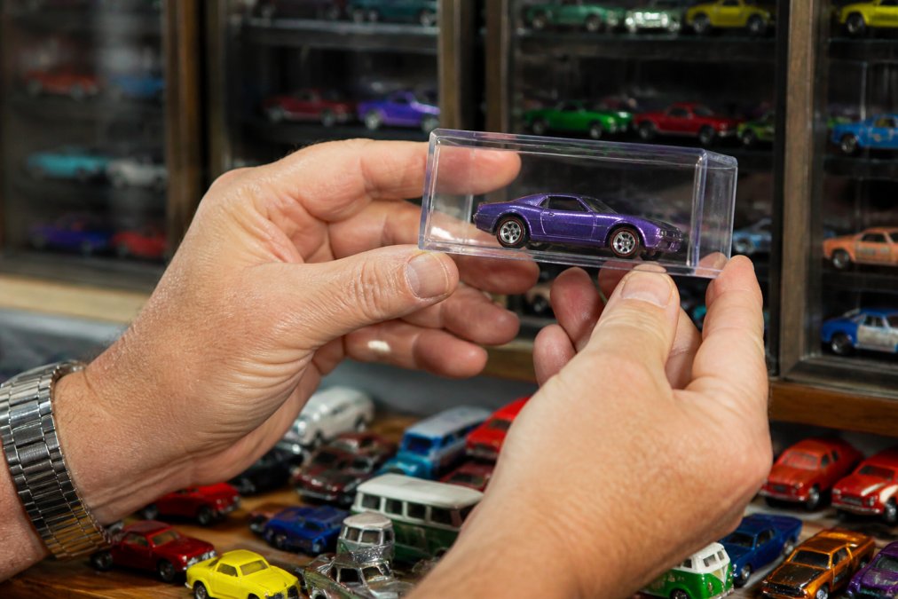 Collector holding a rare Hot Wheels car in a protective case with a display of diecast models in the background