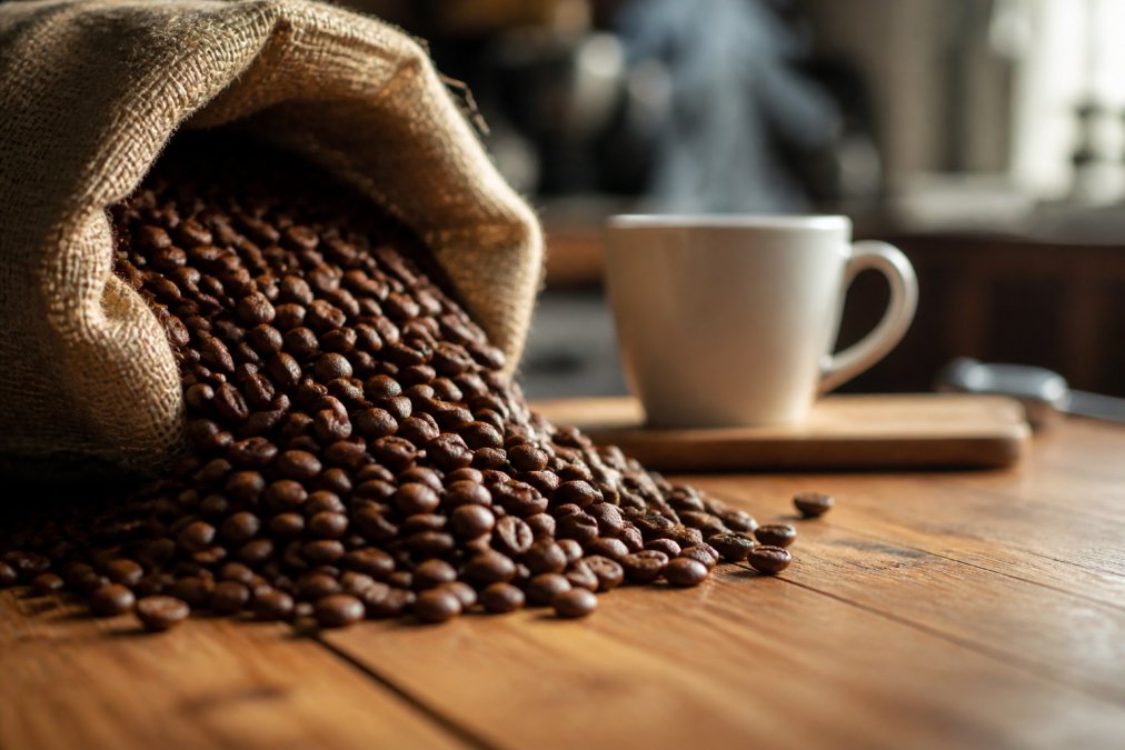 Close-up of roasted coffee beans being poured from a burlap sack onto a wooden counter, with a steaming mug of coffee blurred in the background