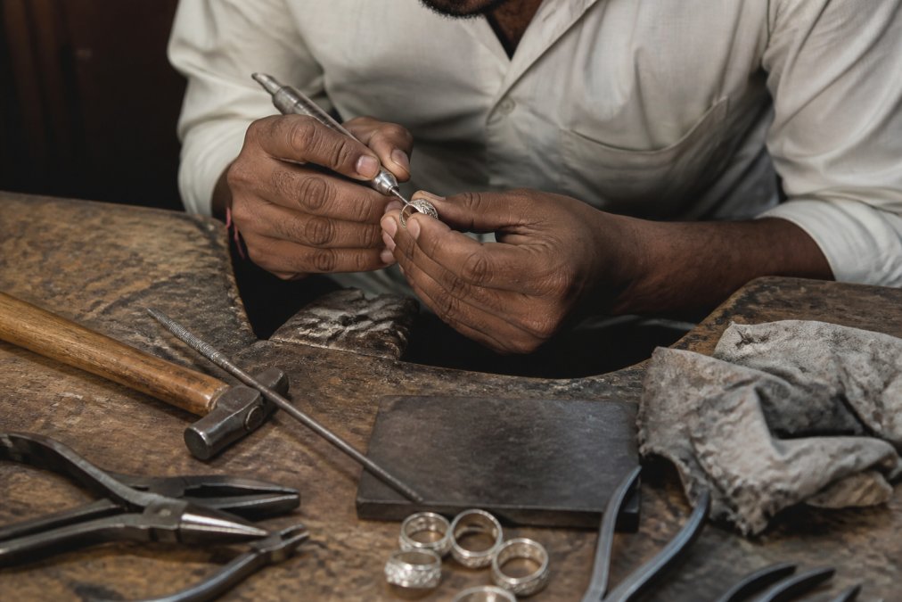 Close-up of skilled artisan hands working on a sterling silver ring with jeweler's tools, files, pliers, and polishing cloths, showing detailed craftsmanship and precision in a workshop setting