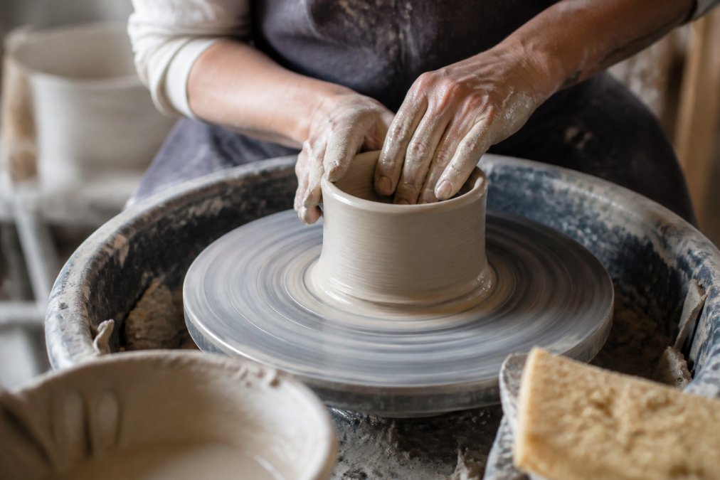 Ceramic artist's hands shaping clay on a spinning pottery wheel, fingers positioned precisely to create even walls, with clay dust visible in soft studio lighting