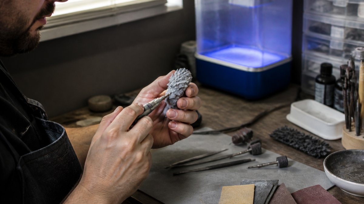 Close-up of hands carefully sanding and finishing a small 3D-printed resin figurine on a workbench, with fine tools, sandpaper, and a curing station visible in a small studio setting