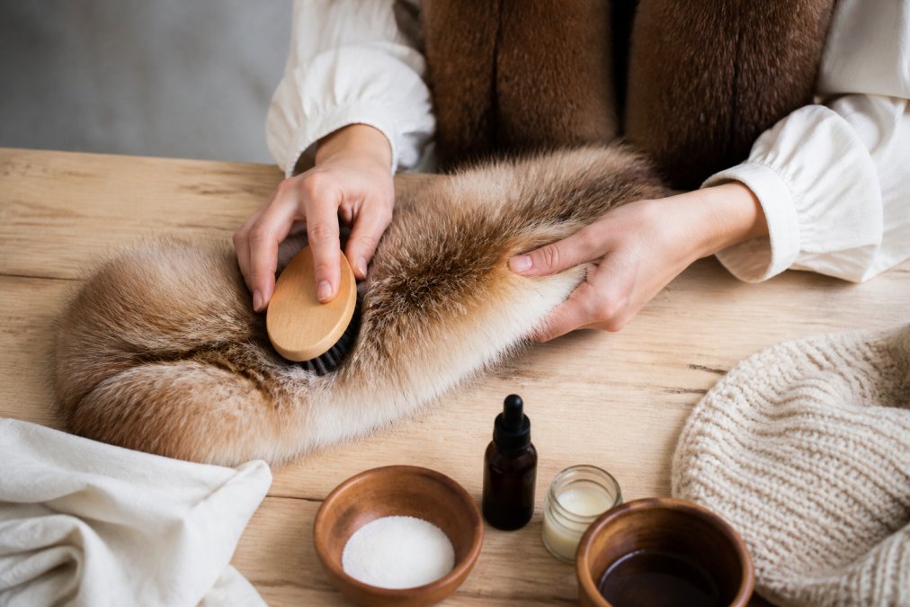 Hands gently brushing and cleaning a natural fur scarf with a soft-bristled brush on a light wooden surface, with care products and tools arranged nearby