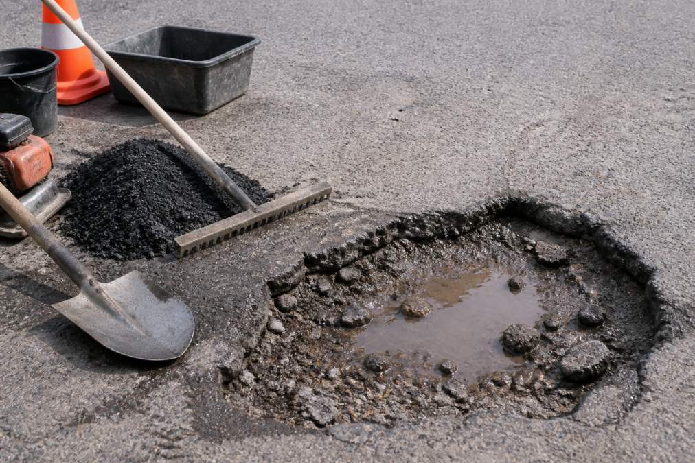 Close-up of a deep pothole in asphalt with visible water pooling, jagged edges, loose aggregate, and exposed subgrade material