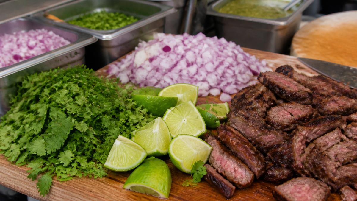 Close-up of vibrant fresh taco ingredients arranged on a wooden counter: bright green cilantro, diced red onions, lime wedges, and charred carne asada with visible grill marks